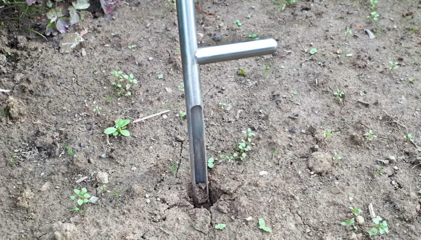 Ein Werkzeug zum Bodenproben ziehen steckt im trockenen Erdreich. Kleine grüne Pflänzchen sprießen zwischen den Rissen im Boden.