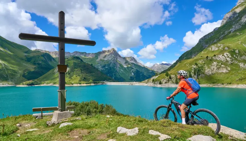 Ciclista en el Alpe Adria Radweg, junto a un lago turquesa y montañas verdes.