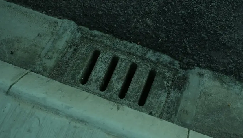 Betonierter Abfluss mit Schlitzen, daneben Asphalt. Der Abfluss erinnert an einen stinkenden revisionsschacht im keller.