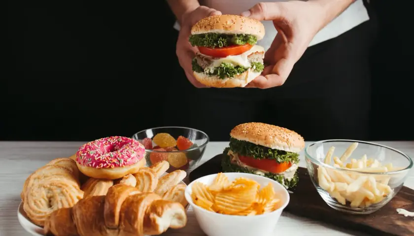 Ręce trzymają burgera, obok talerz z croissantami, pączkiem i ciastkami, miseczki z żelkami, frytkami i chipsami. Czy cheat day przyspiesza metabolizm?