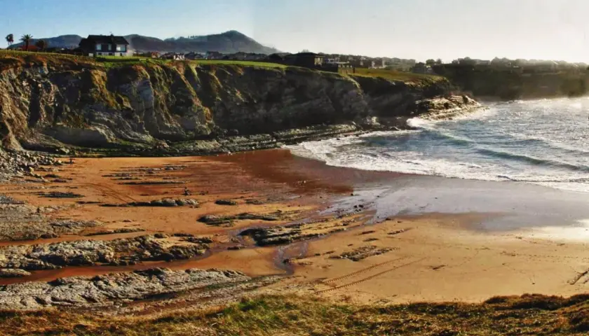 La playa de Portio, con sus acantilados rocosos y arena dorada, se baña en la luz del sol. Las olas rompen suavemente en la costa.
