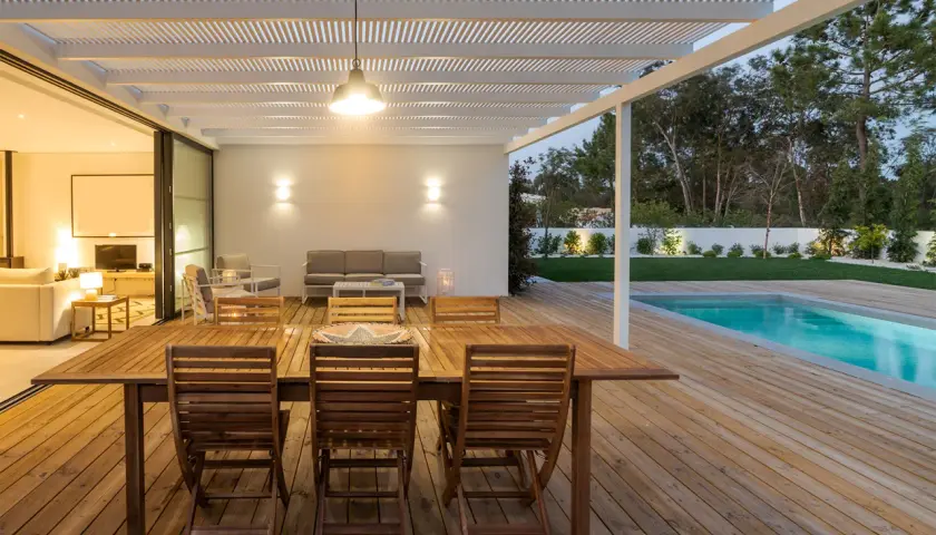 Table et chaises en bois huilé sur une terrasse en bois, avec piscine et jardin en arrière-plan.