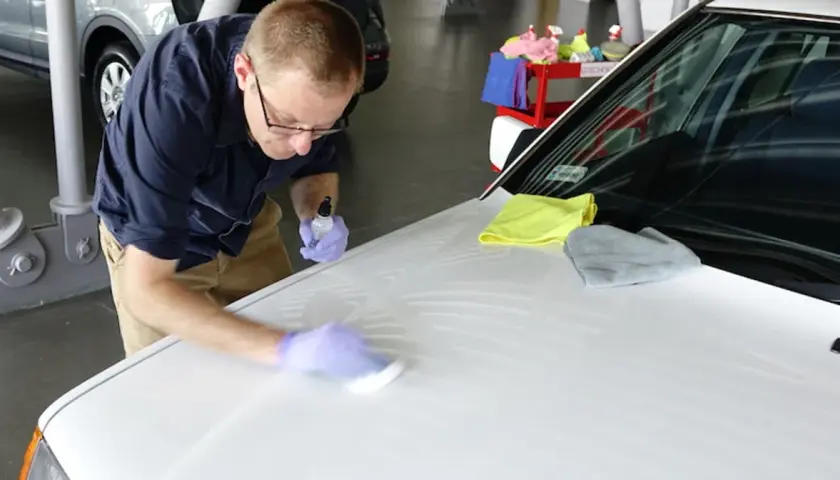 Mężczyzna w rękawiczkach nakłada powłokę ceramiczną na maskę białego samochodu, używając aplikatora i buteleczki z płynem.