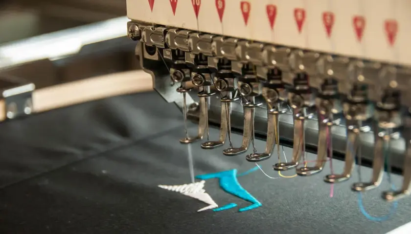 Machine à broder en action, créant un motif coloré sur un tissu noir. La **direct to garment printing** permet des personnalisations uniques.