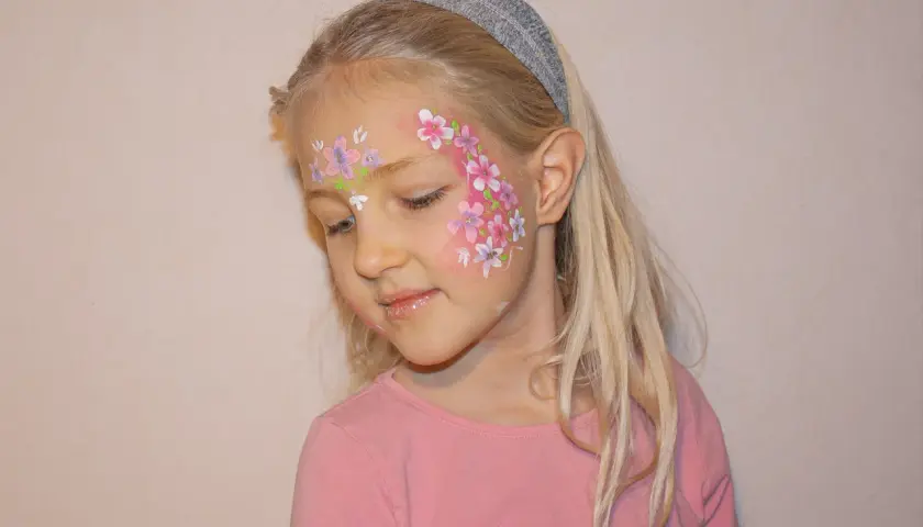 Ein Mädchen mit blonden Haaren und einem Stirnband, das ein Blumenmuster auf der Wange trägt. Das Stern schminken Kinder ist hier als zarte Blüten umgesetzt.