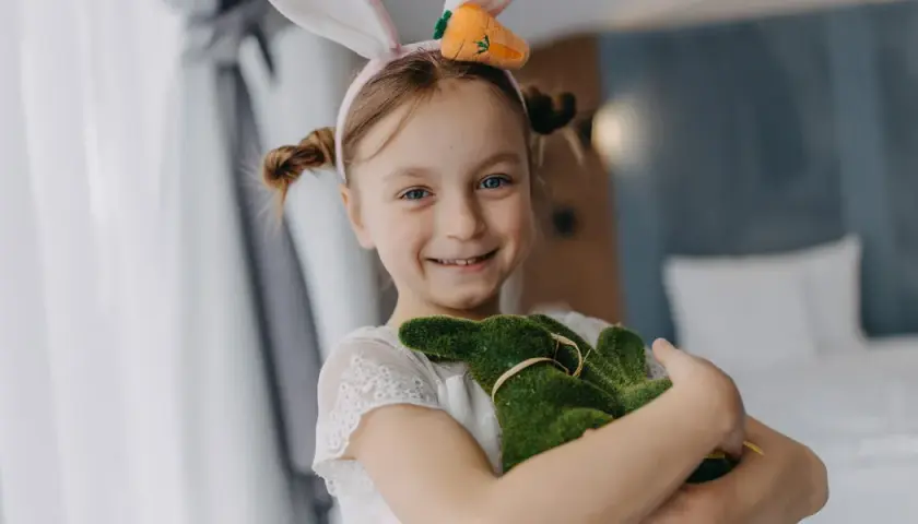 Uśmiechnięta dziewczynka w króliczych uszkach przytula zielonego zajączka. Wesoła wielkanoc w hotelu.