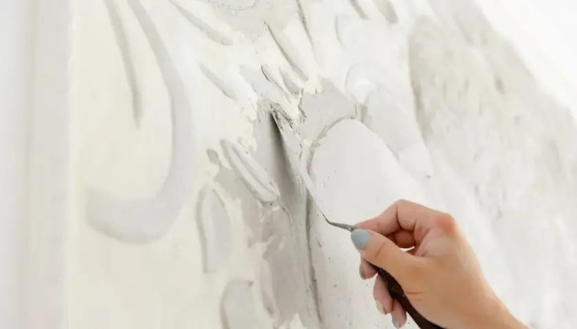 Mano che lavora con uno strumento su uno stucco murale in rilievo, creando un'opera d'arte tridimensionale.