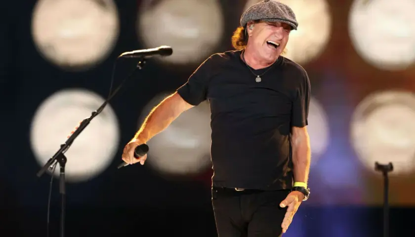 Brian Johnson w czapce i czarnej koszulce śpiewa do mikrofonu na scenie, rozbawiony.