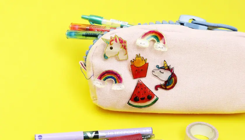 Bunte Schrumpffolie Ideen: Einhorn, Regenbogen, Pommes & Wassermelone zieren eine Federtasche. Stifte & Klebeband liegen daneben.