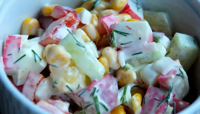 Pyszna sałatka z surimi, kukurydzy, ogórka i jajka, polana kremowym sosem i posypana koperkiem.