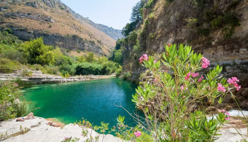 A serene pool of turquoise water reflects the rugged beauty of Sicily nature. Two sunbathers relax on the rocky shore, surrounded by lush greenery and blooming pink oleander flowers.