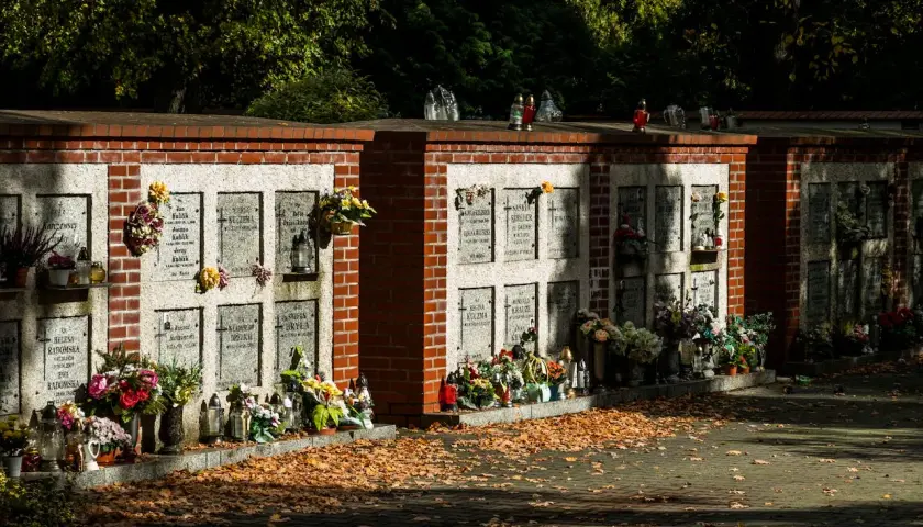 Cmentarne kolumbarium z rzędami nisz na urny, ozdobione kwiatami i zniczami. Jesienne liście pokrywają ścieżkę.