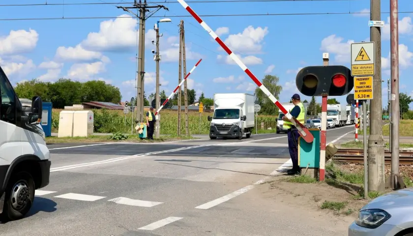 Wjazd na przejazd kolejowy. Zapory opuszczone, czerwone światło sygnalizatora. Pracownik w kamizelce odblaskowej pilnuje ruchu.