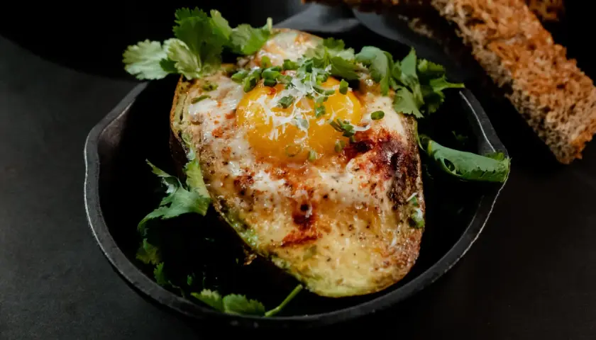 Un avocat oeuf au four, garni de coriandre fraîche et de ciboule, servi dans une poêle en fonte avec du pain grillé.