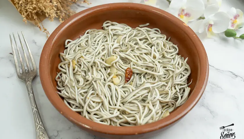 Un cuenco de barro con gulas al ajillo, ajo y guindilla. Un tenedor plateado a la izquierda y flores blancas al fondo.