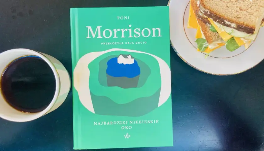 Zielona okładka książki Toni Morrison "Najbardziej niebieskie oko" leży obok filiżanki kawy i kanapki.