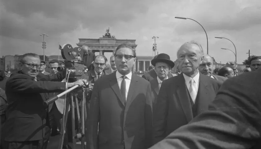 Männer in Anzügen, eine Filmkamera und das Brandenburger Tor im Hintergrund. Ein Mann hält eine "Stalin-Note" in der Hand.