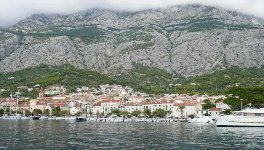Widok na miasto Makarska z górami w tle. W porcie cumują liczne łodzie i jachty. Choć nie ma tu lotniska, widok zapiera dech w piersiach.