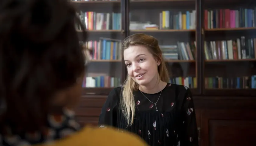 Kobieta z blond włosami, w czarnej bluzce, rozmawia z kimś, kto siedzi tyłem. Za nią regały z książkami. Może to być przykład odwróconej psychologii w związku.