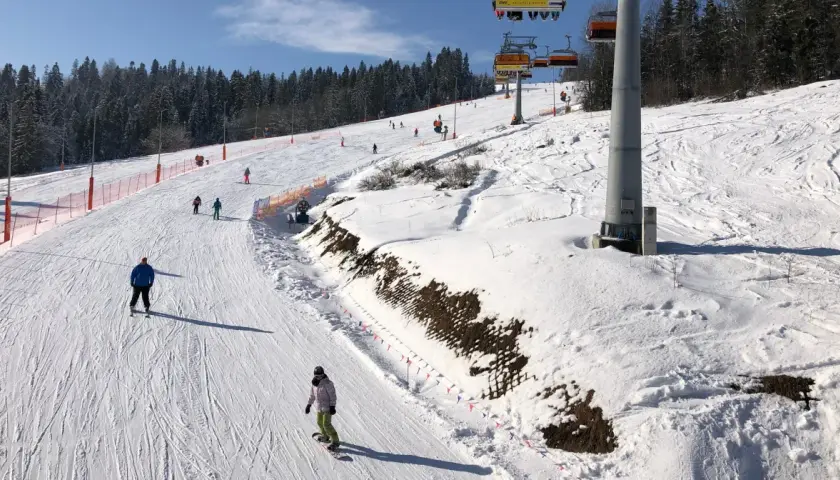 Narciarze i snowboardziści na ośnieżonym stoku z wyciągiem krzesełkowym w tle.