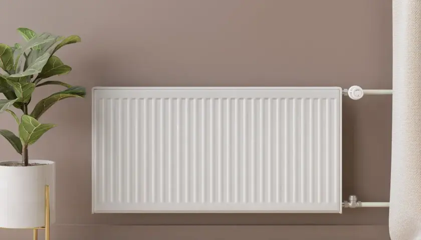 Radiador blanco de panel, un ejemplo de tipos de radiadores de agua. A su lado, una planta en maceta y una cortina.