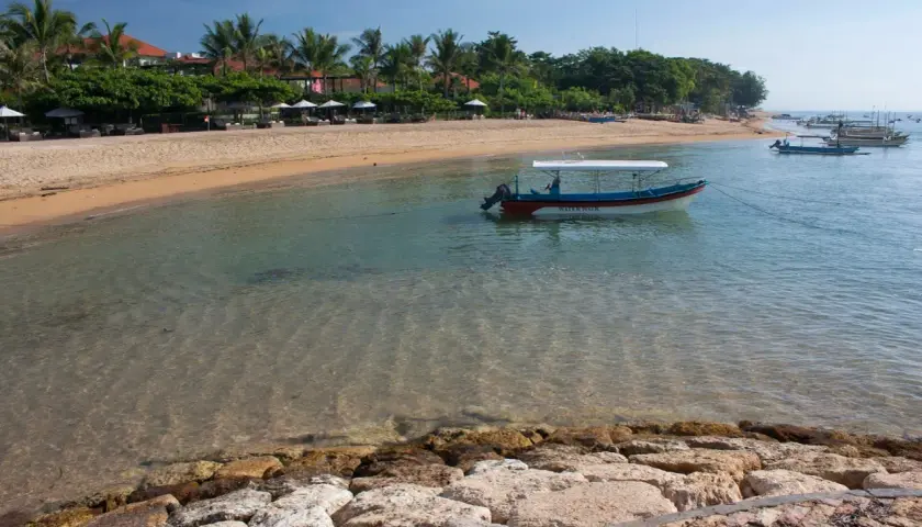 Spokojna zatoka w Sanur z łodzią "Water Walk" kołyszącą się na turkusowej wodzie. Piękna plaża z palmami i leżakami w tle.