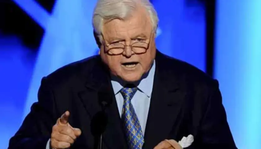 Ein älterer Mann mit Brille und grauem Haar spricht in ein Mikrofon. Er trägt einen dunklen Anzug mit einer blauen Krawatte. Seine Hand ist erhoben, als würde er eine Rede halten, die an die Kennedy-Krankheit erinnert.