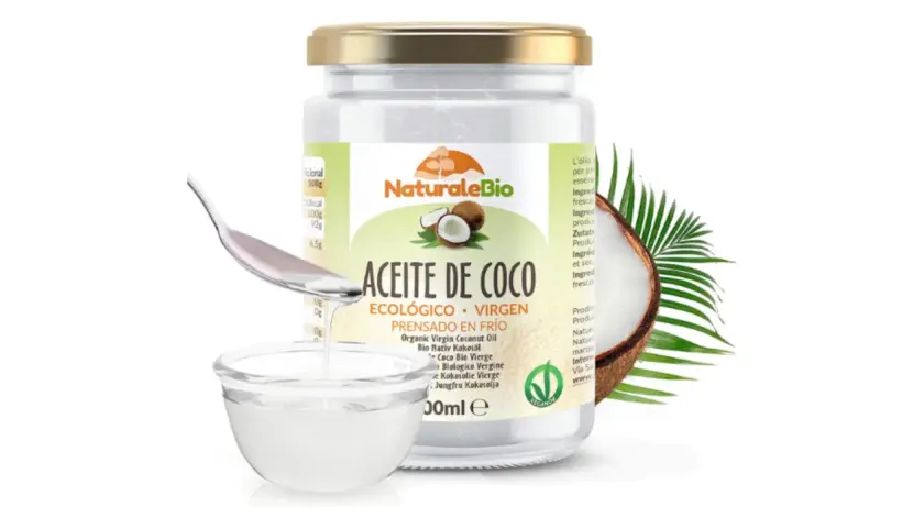Un frasco de aceite de coco ecológico y virgen, ideal para el cuidado del pelo. Una cuchara vierte el aceite en un cuenco.