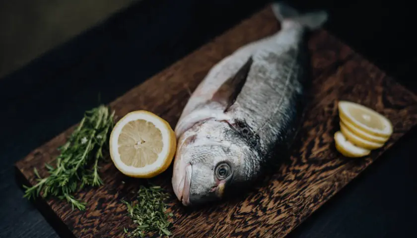Un beau daurade prêt à être grillé, accompagné de citron frais et de romarin sur une planche en bois.