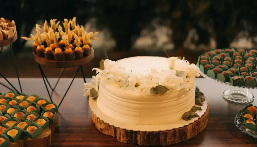Biały tort komunijny ozdobiony białymi kwiatami i zielonymi liśćmi. Obok słodkości, które mogą być inspiracją, jakie kwiaty na tort komunijny.