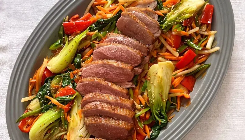 Skivad confiterad anka serveras med en färgstark wok av grönsaker som pak choi, morötter och paprika, toppad med sesamfrön.