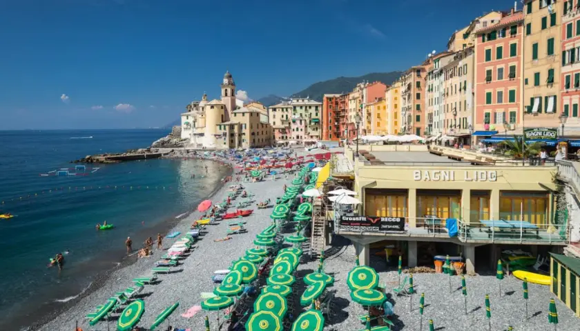 Plage animée à Camogli, Ligurie, avec des parasols verts et jaunes, des bâtiments colorés et la mer bleue.