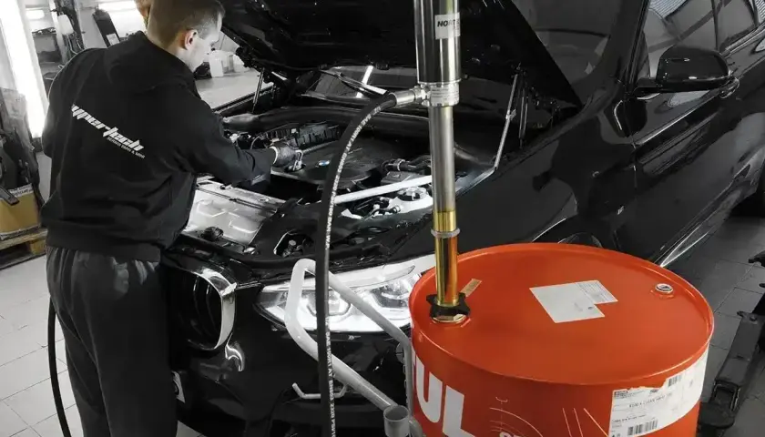 Mechanik wymienia olej w czarnym BMW. Obok stoi duża, pomarańczowa beczka z olejem. Pamiętaj, co ile wymiana oleju jest kluczowa dla silnika.