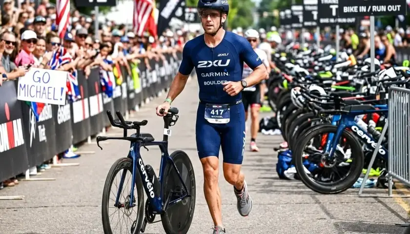 Zawodnik w stroju do triathlonu olimpijskiego biegnie z rowerem obok rzędu rowerów. Tłum kibicuje, trzymając transparent "GO MICHAEL!".