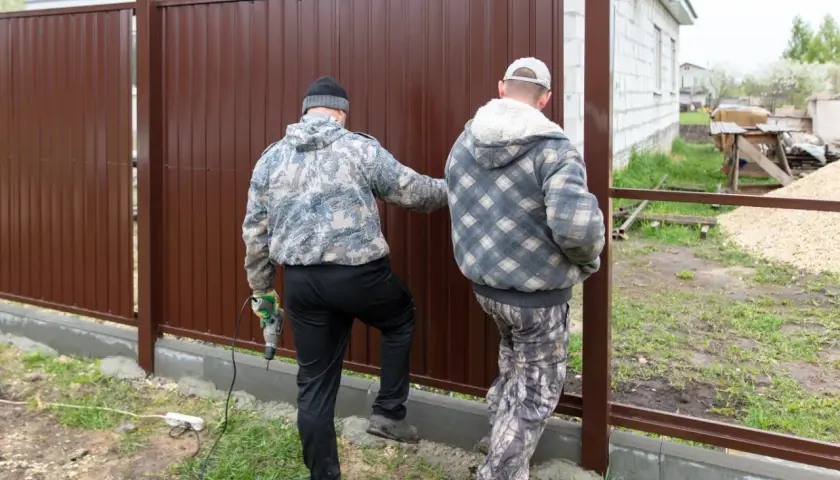 Dwóch mężczyzn montuje brązowe ogrodzenie. Jeden z nich trzyma wiertarkę. Ogrodzenie niezgodne z granicą działki, budowa w toku.
