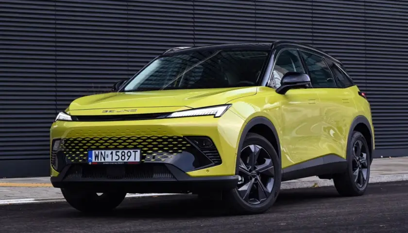 Jasnozielony BAIC 5 z czarnym dachem, nowoczesnym grillem i LED-owymi światłami, zaparkowany przed ciemną, falistą ścianą.
