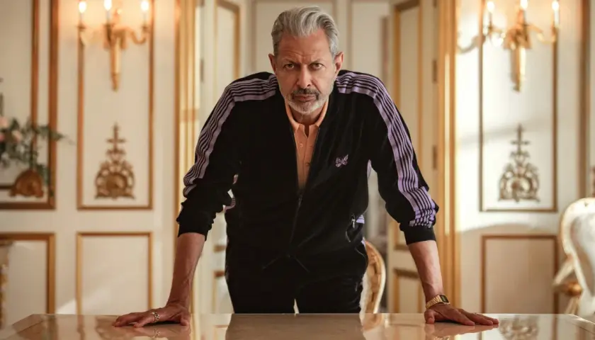 Jeff Goldblum w dresie z fioletowymi pasami, jak z jego filmów, opiera się o stół w eleganckim wnętrzu.