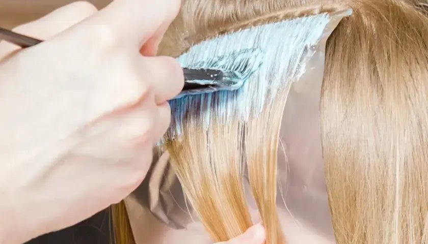 Dłoń z pędzlem nakłada jasnoniebieski rozjaśniacz na pasma blond włosów. Czas ekspozycji jest kluczowy, by uzyskać pożądany efekt.