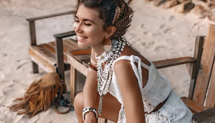 Uśmiechnięta kobieta w stylu boho, ozdobiona muszlami i piórami, siedzi na plaży.