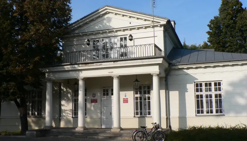 Pałac Potulickich w Pruszkowie, z kolumnowym portykiem i balkonem, otoczony zielenią. Dwa rowery stoją przed wejściem.
