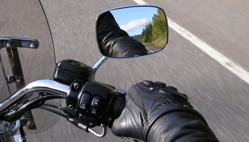 En la carretera, un motorista con guantes de cuero revisa el único espejo de su motocicleta, reflejando el cielo y los árboles.