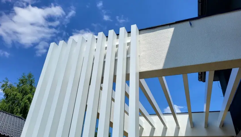 Nowoczesna pergola z białymi, pionowymi lamelami, wykonana z **belki aluminiowe**. Konstrukcja tworzy ciekawy wzór na tle błękitnego nieba z chmurami.