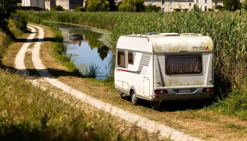 Caravane stationnée au bord d'un canal, dans le Marais Poitevin. Un chemin de terre serpente à travers l'herbe haute.