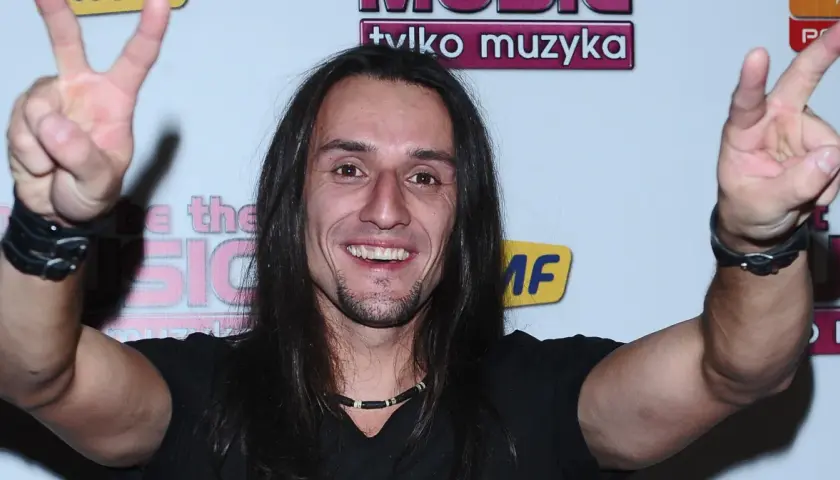 Tomasz Kowalski muzyk z długimi włosami, uśmiechnięty, pokazuje znak pokoju obiema rękami.