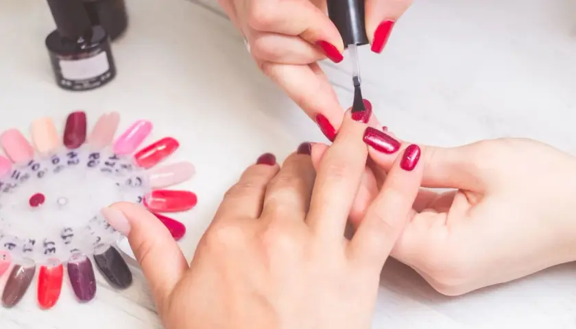 Krok po kroku: jak zrobić manicure. Dłonie w trakcie malowania paznokci na czerwono, obok paleta kolorów lakierów.