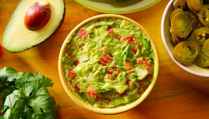 Miseczka guacamole z awokado, pomidorami i cebulą, obok połówka awokado i plasterki jalapeño.