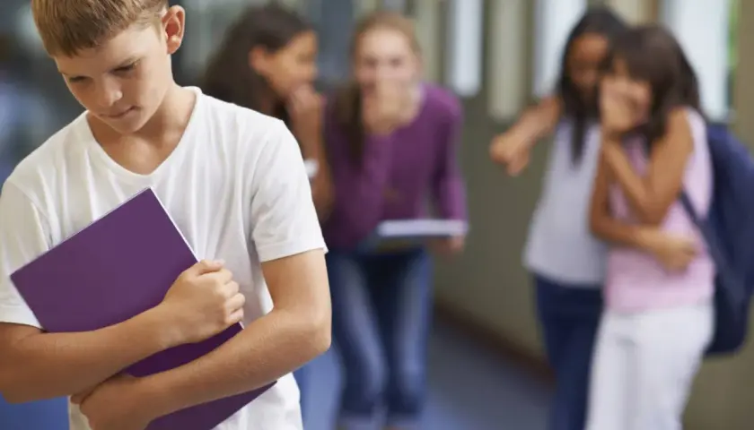 Ragazzo solo con quaderno viola, mentre altri bambini ridono e indicano. Sembra avere problemi con maestre elementari.