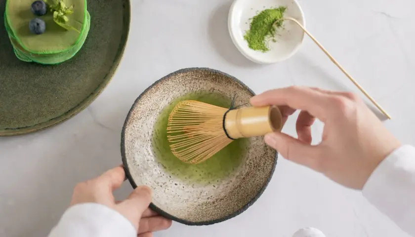 Matcha-Zeit: Hände bereiten grünen Tee mit einem Chasen zu. Daneben ein Stapel grüner Pfannkuchen mit Blaubeeren.
