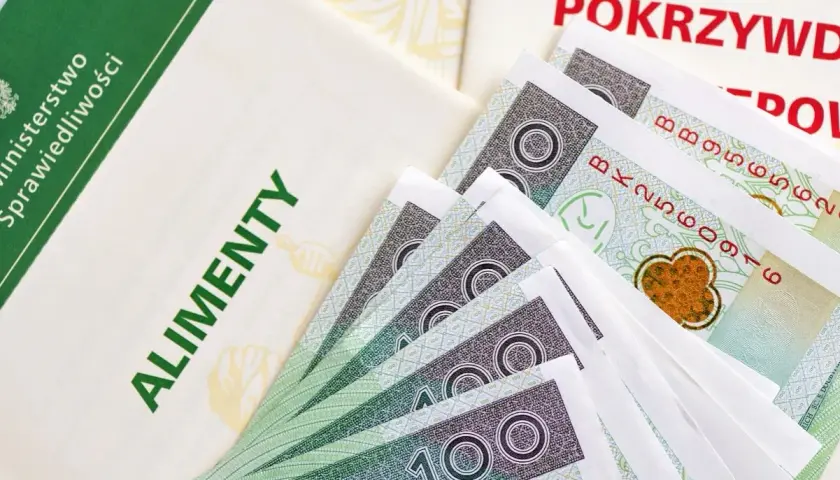 Dokumenty i banknoty 100 zł. Widoczne napisy "ALIMENTY" i "POKRZYWDZONY". Symbolizuje to, jak komornik przelewa alimenty.