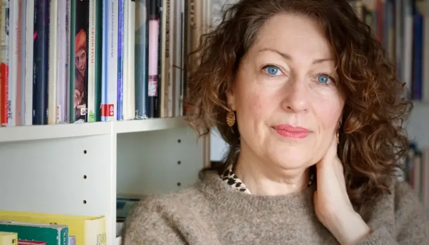 Elisabeth Åsbrink, med blå ögon och lockigt hår, sitter framför en bokhylla. Hon verkar lite sjuk, men hennes blick är klar.
